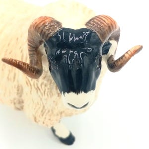 May include: A ceramic figurine of a white sheep with black and brown horns. The sheep has a black face with a white nose and mouth.