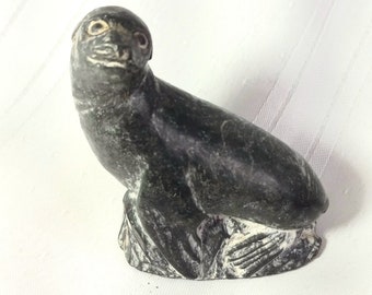 May include: A black soapstone carving of a seal. The seal is sitting on its hind legs with its front flippers tucked in. The carving is detailed with the seal's eyes, nose, and mouth.