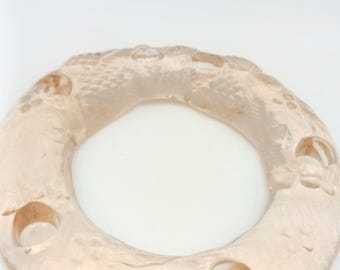 May include: A circular, light peach-colored object with a textured surface and three evenly spaced holes. The object has a ring shape and appears to be made of a translucent material. The design includes subtle patterns and raised details.