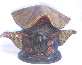 May include: A dark-colored ceramic sculpture featuring a wide, flat, tan-colored top piece resembling a hat. The central body has a textured surface with reddish-brown accents and two pointed, wing-like protrusions extending from the sides.