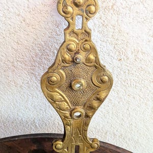 May include: Ornate, antique brass door pull with a decorative, symmetrical design. The brass hardware features a series of keyhole openings and circular accents. The piece is set against a textured, off-white wall and a dark wood surface.