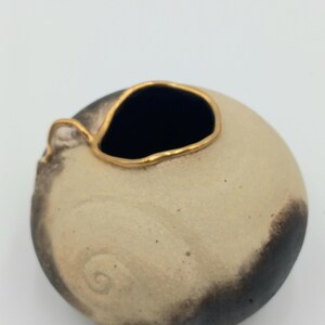 May include: A small, round ceramic vase with a dark interior and a gold-colored rim. The vase has a light beige body with dark brown accents and a small handle. The design includes a spiral detail on the side.