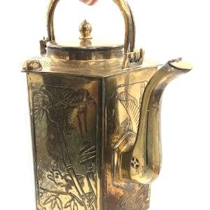 May include: A vintage brass teapot with a square body and a curved spout. The teapot features an ornate handle and lid, with etched bamboo and bird designs on the sides. The teapot is held by a hand.