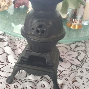 May include: A black cast iron miniature stove with a round body and a small chimney. The stove has a door and sits on four legs. The stove is on a white lace tablecloth.