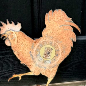 May include: A rustic, orange metal rooster cutout with a circular design on its body. The design features a jar illustration and the text "Warner's Baking Powder". The rooster has a distressed, vintage appearance.