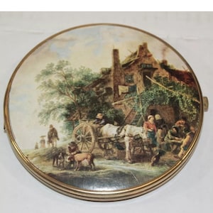 May include: A round, gold-toned compact mirror with a painted scene of a horse-drawn cart and people in a village setting. The scene is painted in muted tones of brown, green, and blue.