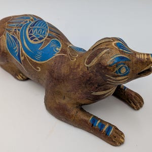 May include: A wooden sculpture of an animal, possibly a fox or dog, in a lying down position. The sculpture is brown with blue and gold painted details. The blue details include floral and geometric patterns on the back and head.