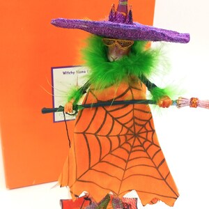 May include: A Halloween-themed figurine of a witch with a purple hat, green boa, and orange dress with a spiderweb design. The figurine holds a broom and wears glittery green shoes. The box behind the figurine has the word "Krinkles".