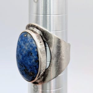 May include: A silver ring featuring a large, oval-shaped, deep blue lapis lazuli stone. The stone is set in a bezel setting, and the ring has a wide, textured band. The ring is displayed on a silver cylindrical object.