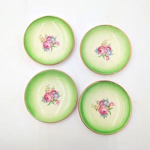 May include: Four small, round plates with a light green rim and a floral design in the center. The flowers are pink, blue, and yellow. The plates have a gold trim and are arranged on a white surface.