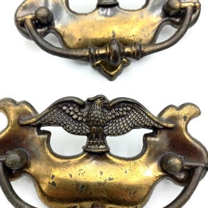 May include: Antique brass drawer pulls with an eagle emblem. The pulls have a curved handle and decorative backplate. The eagle is centered on the backplate with its wings spread. The brass has a vintage, aged appearance.