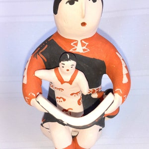 May include: Hand-painted ceramic figurine of a woman holding a child. The woman is wearing a red and black dress with white accents. The child is wearing a red and white dress with black accents. Both figures have black hair and brown eyes.