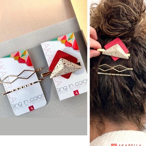Puede incluir: Accesorios para el cabello con diseños geométricos. Una horquilla tiene una base cuadrada roja con un triángulo moteado encima. Otra tiene un diseño en zigzag dorado. El texto "Sing in color SPEAK IN TEXTURE" es visible.