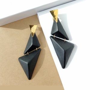 Puede incluir: Un par de pendientes de cuero negro con detalles dorados. Los pendientes tienen forma de triángulos y están superpuestos. Los pendientes están hechos de cuero reciclado y forman parte de una colección de joyería sostenible. "RECYCLED LEATHER JEWELRY" está escrito en texto marrón sobre un fondo blanco.