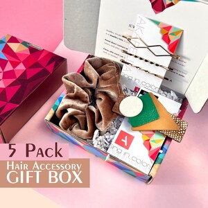 Puede incluir: Una caja de regalo llena de cinco accesorios para el cabello, incluyendo un scrunchie marrón, pinzas para el cabello doradas y pinzas para el cabello de cuero. La caja está decorada con un patrón geométrico colorido y el texto "Sing in colors, Speak in textures".