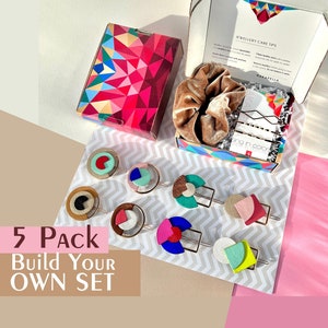 Puede incluir: Un conjunto de cinco pinzas para el pelo de colores con formas geométricas de cuero. Las pinzas están dispuestas sobre una superficie con estampado blanco y gris. El texto "5 Pack Build Your Own Set" es visible en la imagen.