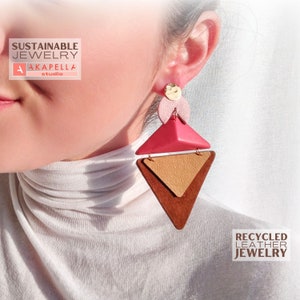 Puede incluir: Un par de pendientes geométricos hechos de cuero reciclado. Los pendientes son marrones, rosas y dorados. Los pendientes tienen forma de triángulo con un triángulo más pequeño en su interior. Los pendientes cuelgan de la oreja de una persona.