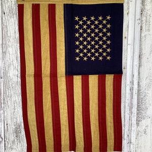 May include: A vintage American flag with red and gold stripes and a blue field of stars. The flag is hanging on a white wooden wall.