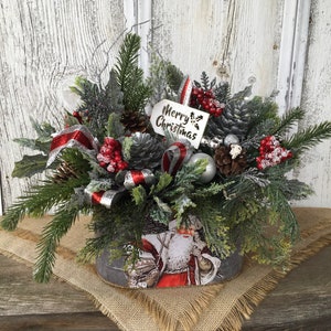 May include: A Christmas centerpiece in a gray metal container with a Santa Claus design. The centerpiece features a variety of greenery, red berries, pine cones, and a silver ribbon. A white tag with the text "Merry Christmas" is attached to the arrangement.