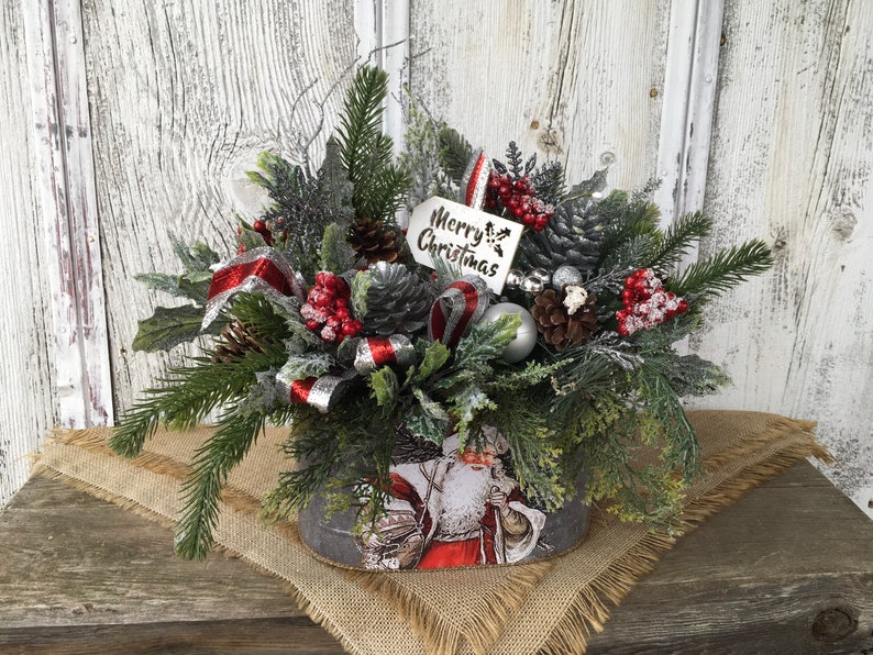 May include: A Christmas centerpiece in a gray metal container with a Santa Claus design. The centerpiece features a variety of greenery, red berries, pine cones, and a silver ribbon. A white tag with the text "Merry Christmas" is attached to the arrangement.