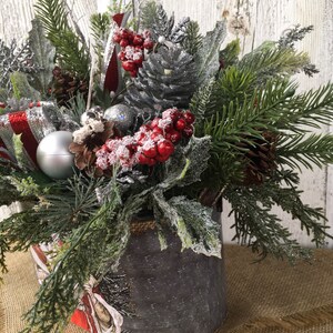 May include: A silver metal container with a burlap rim filled with a festive Christmas arrangement. The arrangement includes pine sprigs, red berries, silver pine cones, and a silver ornament.