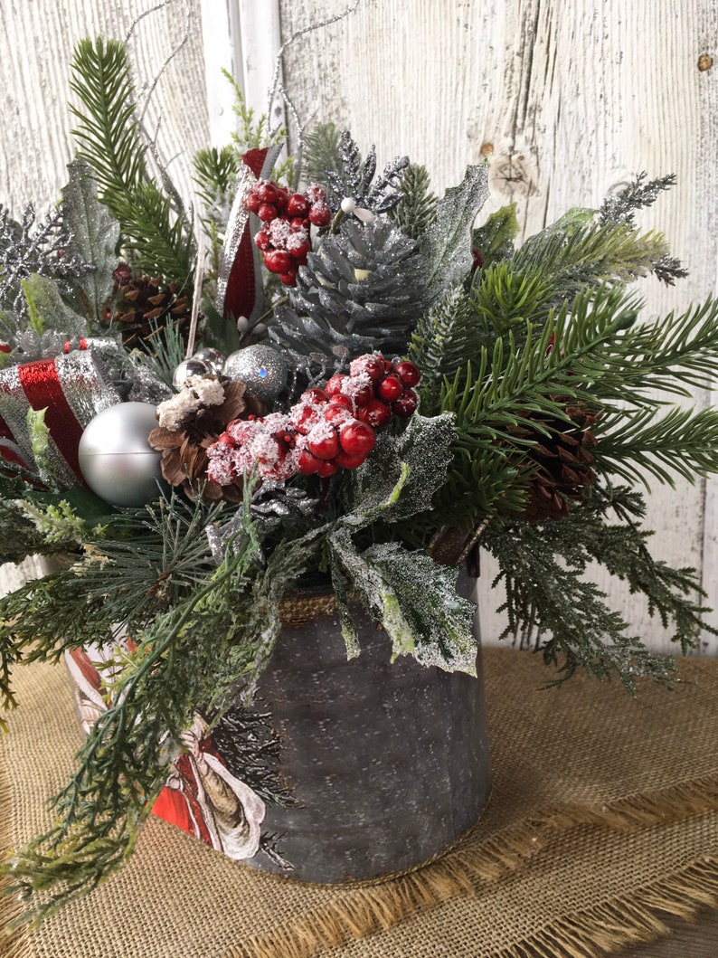May include: A silver metal container with a burlap rim filled with a festive Christmas arrangement. The arrangement includes pine sprigs, red berries, silver pine cones, and a silver ornament.