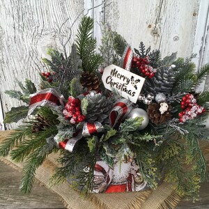 May include: A Christmas centerpiece in a metal container with a Santa Claus design. The centerpiece features a variety of greenery, pine cones, red berries, silver ribbon, and a silver ornament. A white wooden tag with the text "Merry Christmas" is attached to the arrangement.