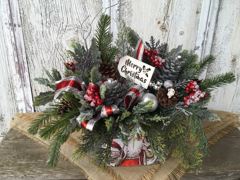 May include: A Christmas centerpiece in a metal container with a Santa Claus design. The centerpiece features a variety of greenery, pine cones, red berries, silver ribbon, and a silver ornament. A white wooden tag with the text "Merry Christmas" is attached to the arrangement.