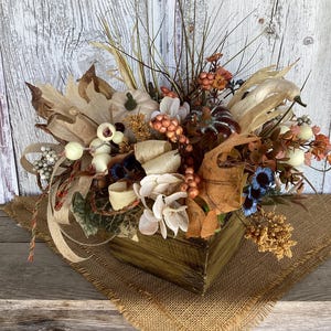 May include: A rustic wooden box overflowing with a fall-themed arrangement. It features miniature pumpkins, dried leaves, berries, and faux flowers in shades of beige, brown, orange, and blue. The arrangement sits on a burlap cloth, creating a warm, autumnal display.