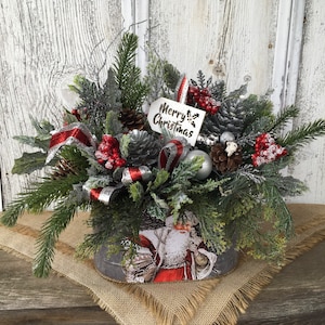 May include: A Christmas centerpiece in a gray metal container with a Santa Claus design. The centerpiece features a variety of greenery, red berries, pine cones, and a silver ribbon. A white tag with the text "Merry Christmas" is attached to the arrangement.