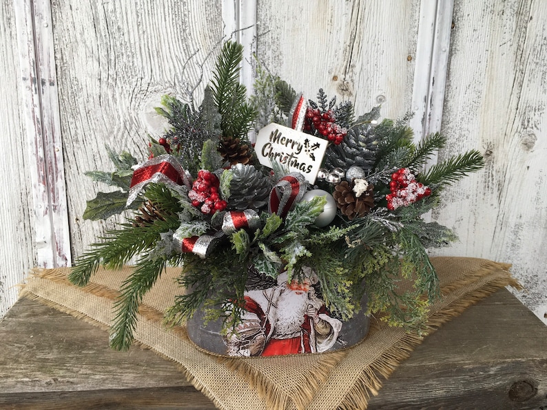 May include: A Christmas centerpiece in a gray metal container with a Santa Claus design. The centerpiece features a variety of greenery, red berries, pine cones, and a silver ribbon. A white tag with the text "Merry Christmas" is attached to the arrangement.