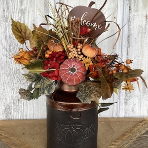 May include: A rustic metal milk can vase with a copper top, decorated with a pumpkin design, filled with a fall floral arrangement featuring red, orange, and yellow flowers, leaves, and berries. The arrangement is topped with a metal pumpkin sign that reads "Welcome".