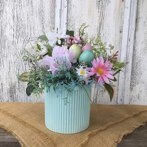 May include: A pastel blue ceramic vase with a ribbed texture, filled with a floral arrangement of pink, white, and green flowers. The vase is decorated with pastel colored Easter eggs.
