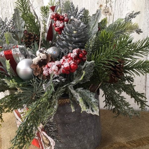 May include: A silver metal container with a burlap rim filled with a festive Christmas arrangement. The arrangement includes pine sprigs, red berries, silver pine cones, and a silver ornament.