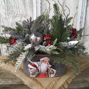 May include: A festive Christmas centerpiece featuring a silver metal container with a vintage Santa Claus image. The container is filled with frosted greenery, red berries, and a silver ornament.