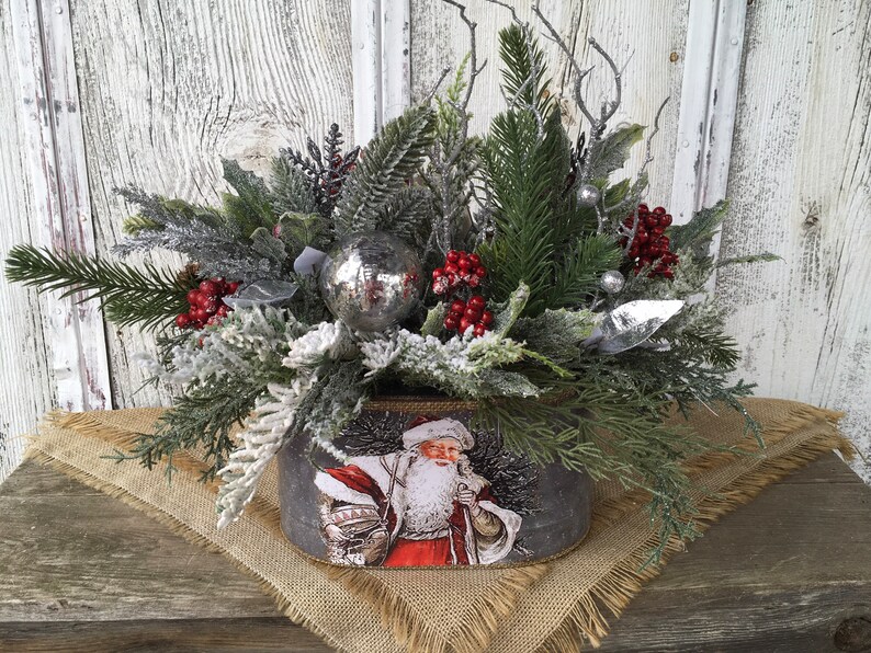 May include: A festive Christmas centerpiece featuring a silver metal container with a vintage Santa Claus image. The container is filled with frosted greenery, red berries, and a silver ornament.