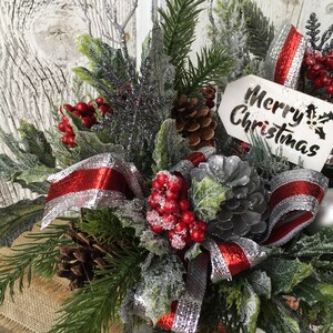 May include: A festive Christmas centerpiece with a red and silver ribbon, pine branches, red berries, pinecones, and a sign that reads "Merry Christmas".