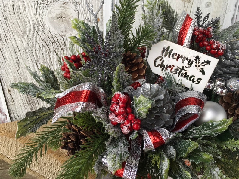 May include: A festive Christmas centerpiece with a red and silver ribbon, pine branches, red berries, pinecones, and a sign that reads "Merry Christmas".