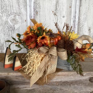 May include: A fall-themed centerpiece in a gray metal container featuring orange sunflowers, a small orange pumpkin, brown pinecones, dried wheat, and a beige burlap bow. Two triangular candy corn-shaped decorations sit to the left.