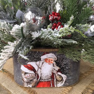 May include: A gray metal planter with a festive Christmas design featuring a vintage Santa Claus illustration. The planter is filled with snowy green pine branches, red berries, and a silver ornament.