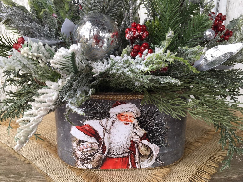 May include: A gray metal planter with a festive Christmas design featuring a vintage Santa Claus illustration. The planter is filled with snowy green pine branches, red berries, and a silver ornament.