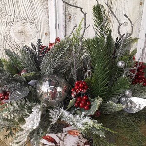 May include: A winter-themed centerpiece with a silver ornament, red berries, and frosted greenery. The centerpiece is in a rustic wooden box with a Santa Claus image.