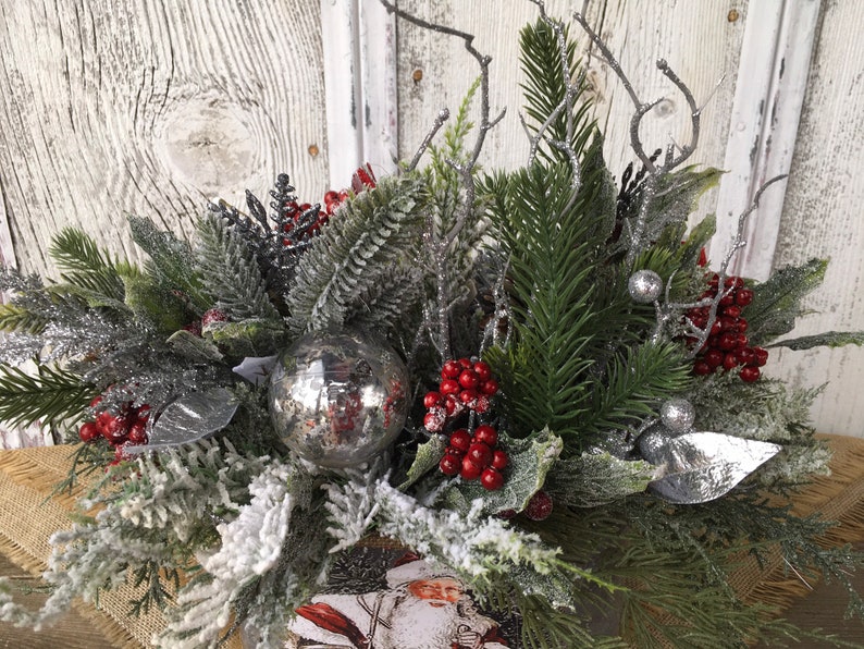 May include: A winter-themed centerpiece with a silver ornament, red berries, and frosted greenery. The centerpiece is in a rustic wooden box with a Santa Claus image.