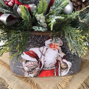 May include: A gray metal planter with a vintage image of Santa Claus carrying a sack of gifts. The planter is decorated with a burlap ribbon and is filled with a festive arrangement of greenery, red berries, and a silver ornament.