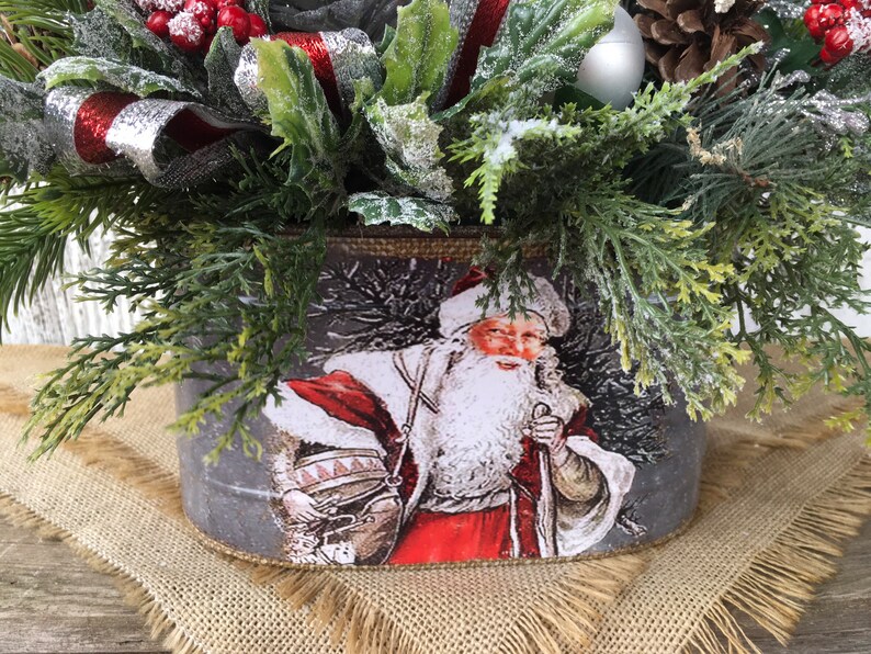 May include: A gray metal planter with a vintage image of Santa Claus carrying a sack of gifts. The planter is decorated with a burlap ribbon and is filled with a festive arrangement of greenery, red berries, and a silver ornament.