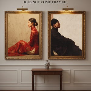 May include: Two framed portraits hang on a wall, each with a gold frame and a warm-toned image. One depicts a person in a red dress, the other a person in black clothing. A small wooden table and a decorative vase are below the portraits. Text above the frames reads "DOES NOT COME FRAMED."