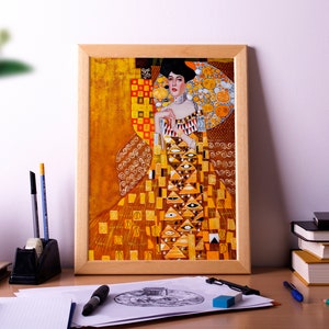 May include: A framed print of a Gustav Klimt painting, featuring a woman in an ornate, gold-toned dress. The artwork has a warm color palette with geometric patterns. The frame is light brown. The print is on a desk with art supplies.