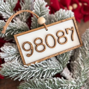 May include: Wooden Christmas ornament with the number 98087 in a brown font on a white background. The ornament has a wooden frame and a natural twine loop with a wooden bead for hanging. The ornament is displayed on a bed of frosted evergreen branches.