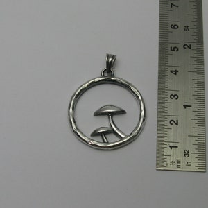 Pode incluir: Colar com pingente de prata com duas silhuetas de cogumelos dentro de um c&iacute;rculo martelado. O pingente mede aproximadamente 3,8 cm de di&acirc;metro.