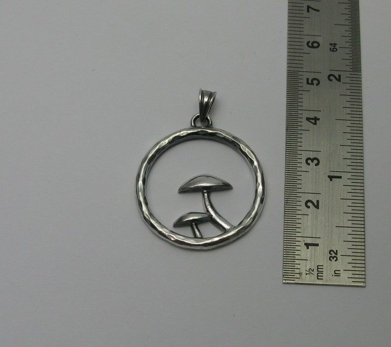 Pode incluir: Colar com pingente de prata com duas silhuetas de cogumelos dentro de um c&iacute;rculo martelado. O pingente mede aproximadamente 3,8 cm de di&acirc;metro.